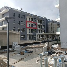 ABC - Wielopiętrowy budynek mieszkalny w trakcie prac wykończeniowych elewacji, z pracownikiem na dachu i fragmentem elewacji zaznaczonym czerwoną ramką, widoczne materiały budowlane na placu budowy.