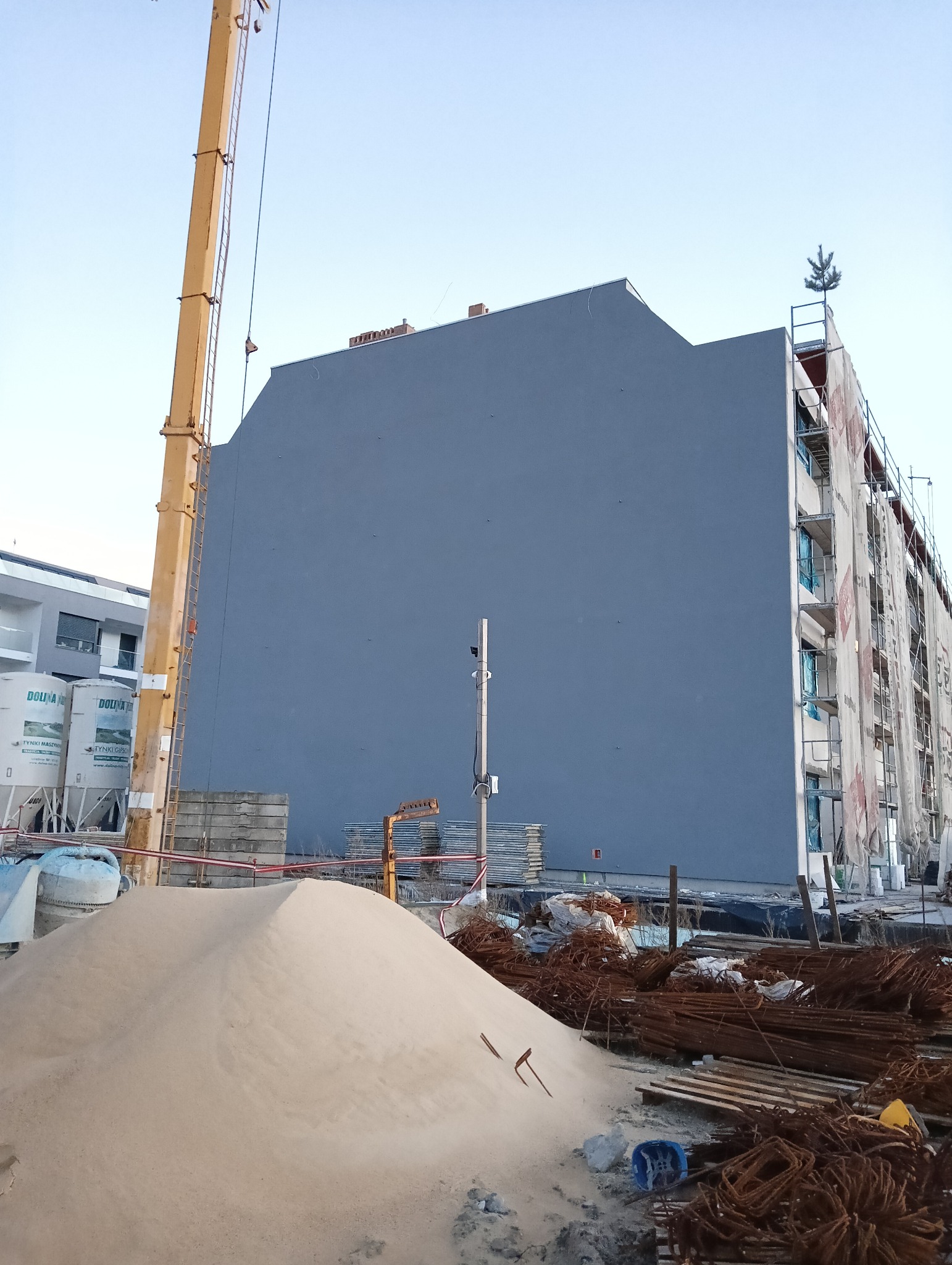 Szara ściana budynku w trakcie prac elewacyjnych, widoczne rusztowania z siatką ochronną, dźwig budowlany, hałda piasku i materiały budowlane na placu budowy.