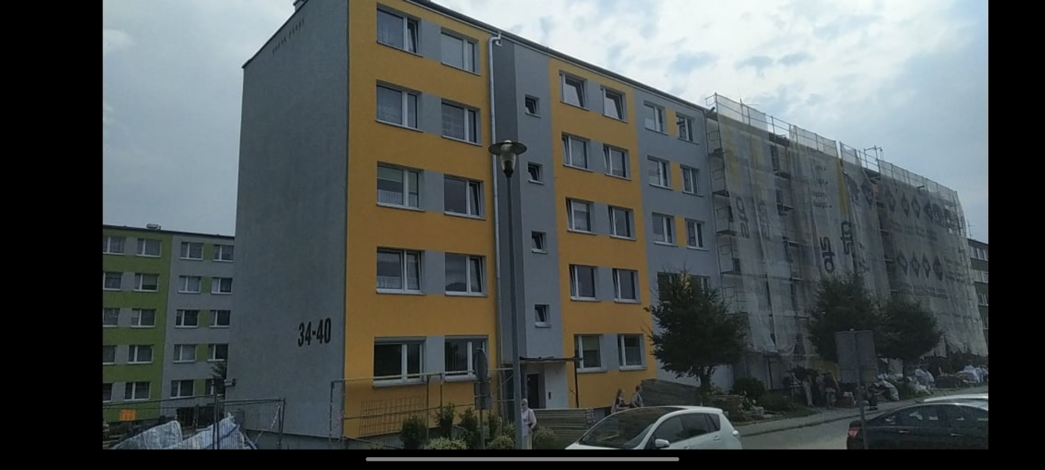 Wielopiętrowy budynek mieszkalny z częściowo odnowioną elewacją w kolorach szarym i żółtym, część budynku w rusztowaniach osłoniętych siatką, sąsiedni budynek w odcieniach zieleni i szarości.