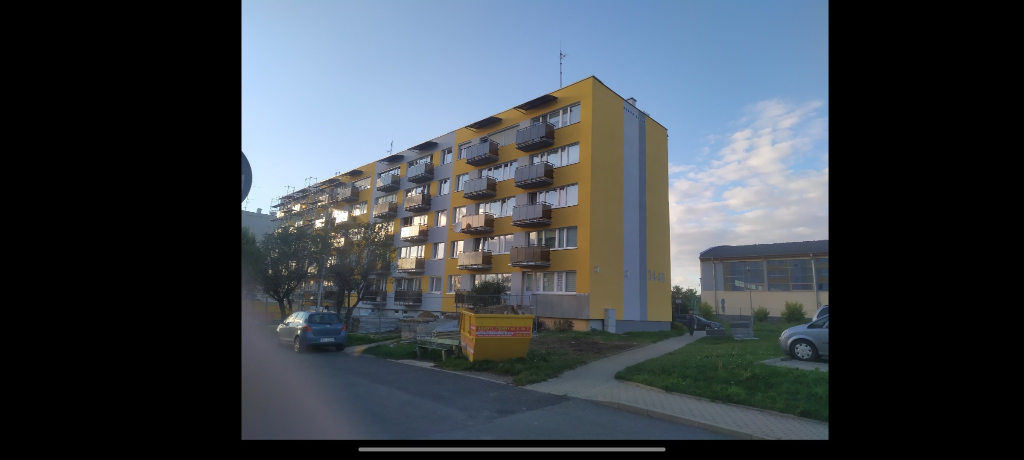 Wielopiętrowy budynek mieszkalny z balkonami, częściowo odnowiony, z żółtą i szarą elewacją, widoczne rusztowania, kontener na odpady budowlane i zaparkowane samochody.