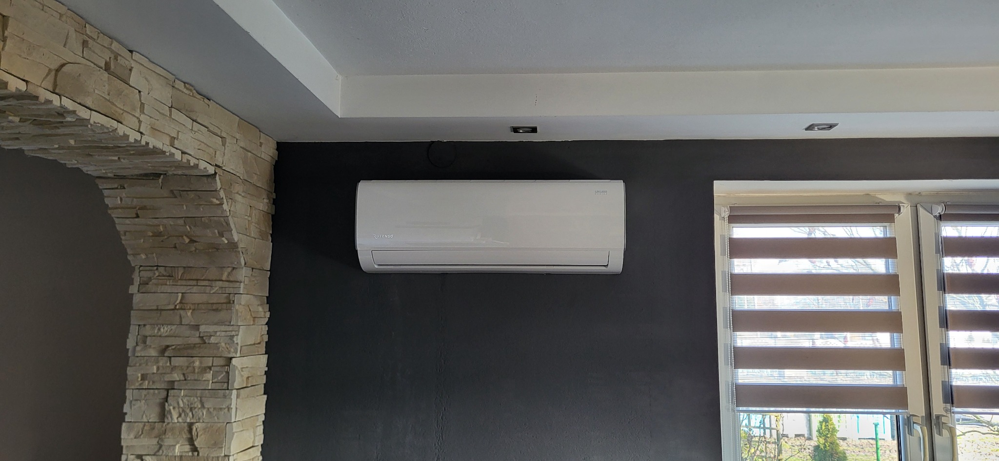 Biały klimatyzator na ciemnoszarej ścianie w pokoju z kamiennym łukiem i oknem z roletami.