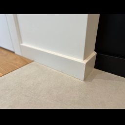 Barthouse Bartosz Waksmundzki - Detal wykończenia: biała listwa przypodłogowa z MDF, łącząca się z podłogą z jasnych płytek. Widoczny fragment drewnianej podłogi i czarna ściana w tle.