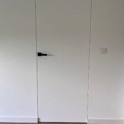 Barthouse Bartosz Waksmundzki - Minimalistyczne wnętrze: białe ściany z ukrytymi drzwiami i czarną klamką, listwa przypodłogowa i panele podłogowe w odcieniach brązu. Nowoczesny design.