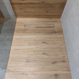 Barthouse Bartosz Waksmundzki - Ukończona kabina prysznicowa z drewnopodobnymi płytkami i odpływem liniowym. Jasne płytki na ścianach. Widok z góry, wnętrze łazienki.