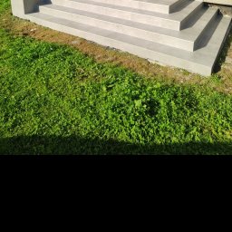 Barthouse Bartosz Waksmundzki - Zewnętrzne schody wyłożone jasnoszarymi płytkami, z widoczną zieloną trawą i fragmentem elewacji budynku. Kąt ujęcia z dołu podkreśla solidność wykonania.