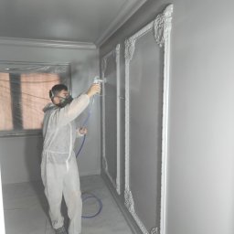 Barthouse Bartosz Waksmundzki - Malowanie natryskowe szarego wnętrza z dekoracyjnymi listwami przez pracownika w kombinezonie ochronnym i masce. Widoczny pistolet natryskowy i wąż.
