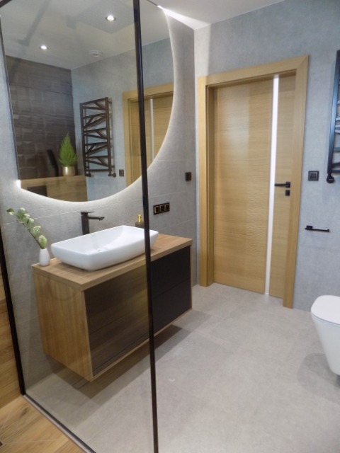 Nowoczesna łazienka z asymetrycznym lustrem, drewnianą szafką pod umywalką i szarą glazurą. Widoczna kabina prysznicowa ze szkła.