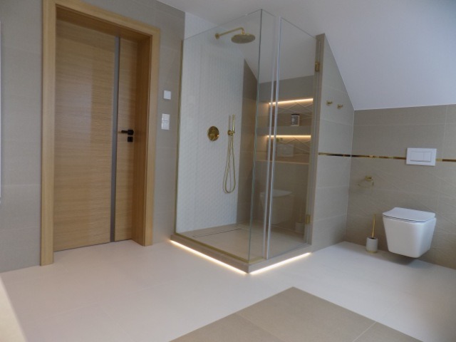 Nowoczesna łazienka z kabiną prysznicową typu walk-in, złotymi akcentami i podświetleniem LED. Minimalistyczny design, beżowe płytki i drewniane drzwi.