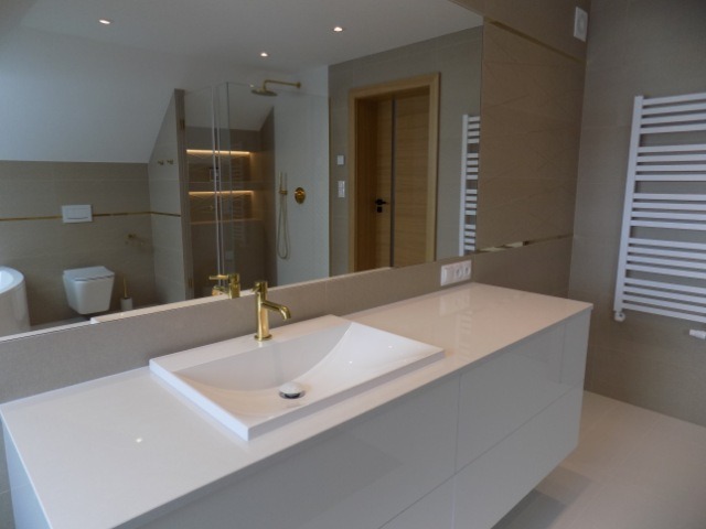 Biała umywalka z baterią w kolorze złota na blacie w eleganckiej łazience z beżowymi płytkami i grzejnikiem drabinkowym. Widok w lustrze na kabinę prysznicową.