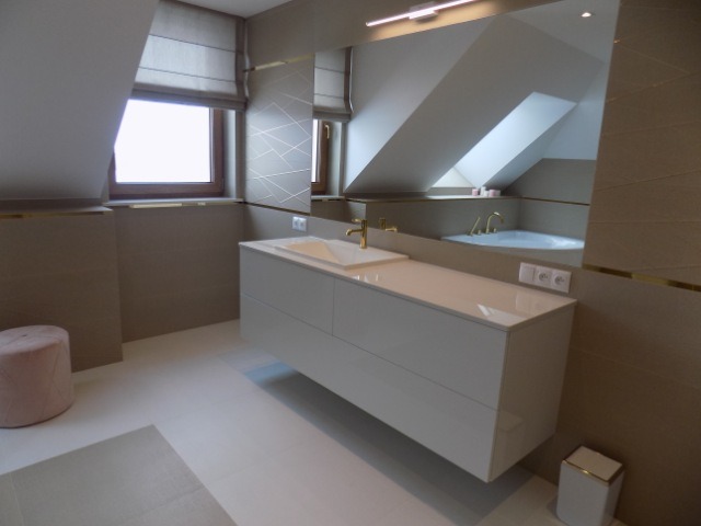 Jasna łazienka na poddaszu z beżowymi ścianami, białymi meblami łazienkowymi i złotymi akcentami. W lustrze odbija się wanna. Nowoczesny design.