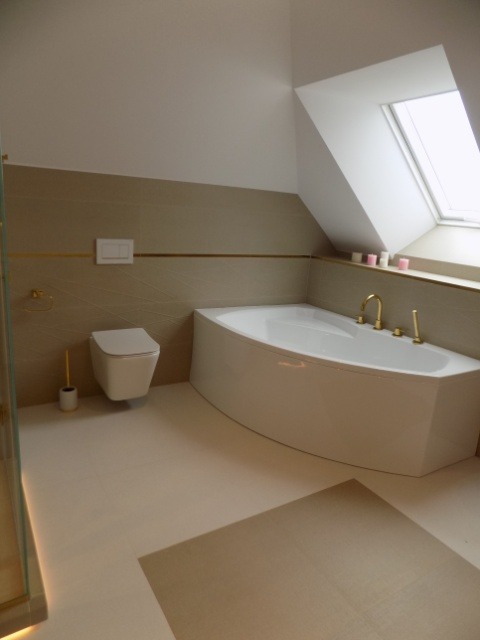 Jasna łazienka na poddaszu z wanną narożną, wiszącą toaletą i oknem dachowym, wykończona beżowymi płytkami z subtelnym złotym akcentem.