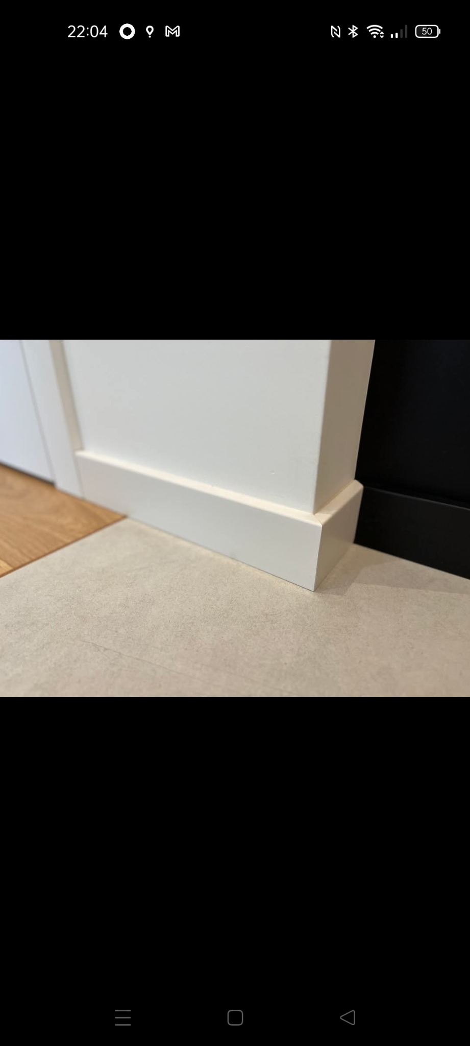 Detal wykończenia: biała listwa przypodłogowa z MDF, łącząca się z podłogą z jasnych płytek. Widoczny fragment drewnianej podłogi i czarna ściana w tle.