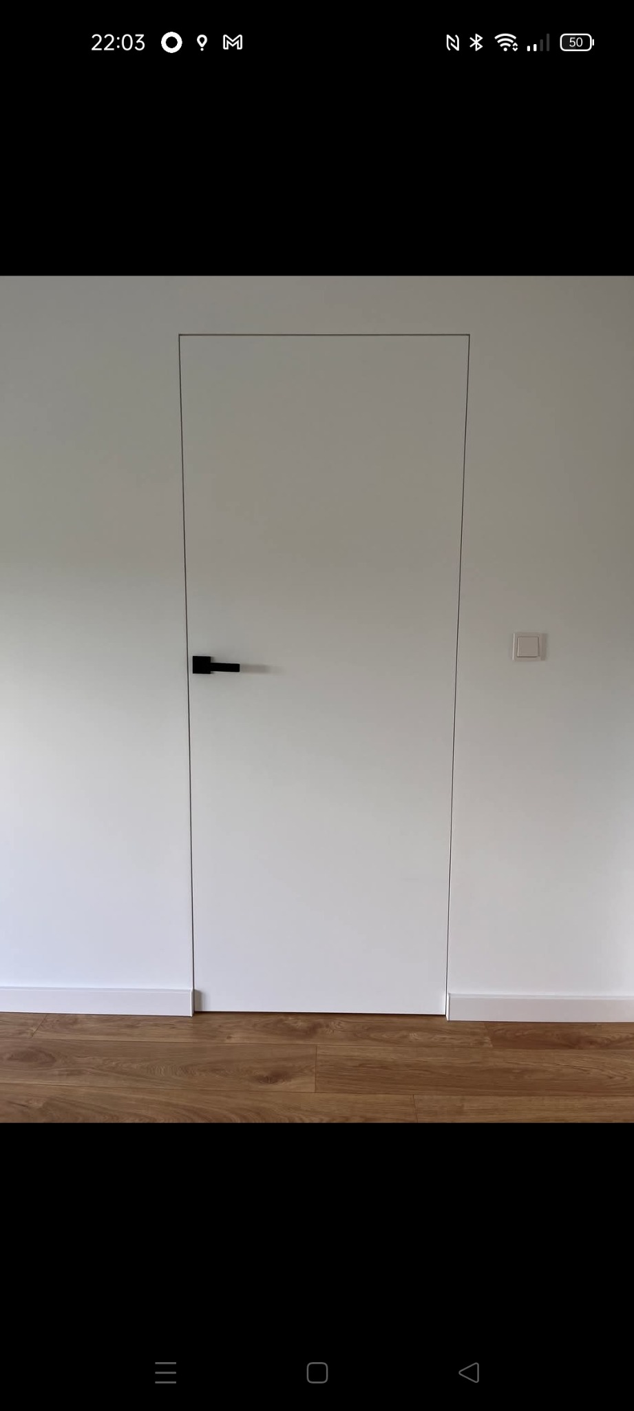 Minimalistyczne wnętrze: białe ściany z ukrytymi drzwiami i czarną klamką, listwa przypodłogowa i panele podłogowe w odcieniach brązu. Nowoczesny design.