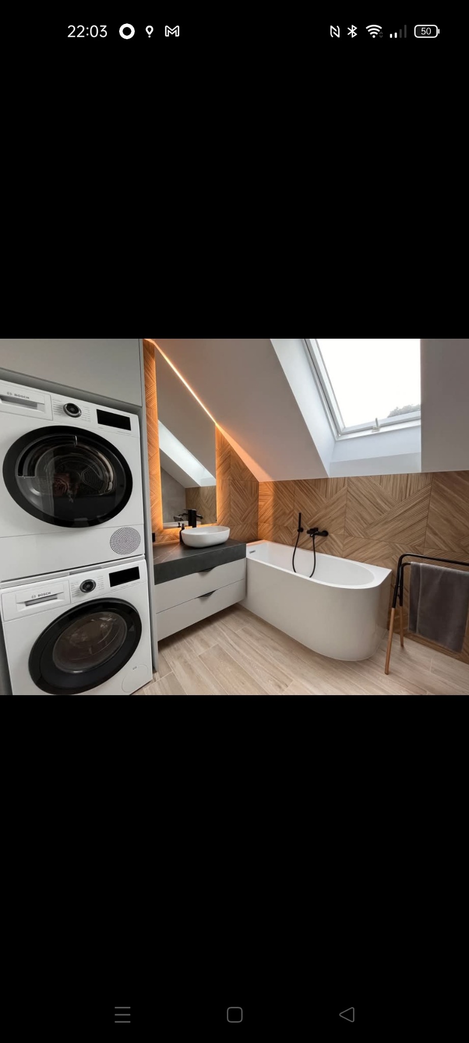 Nowoczesna łazienka na poddaszu z drewnianą mozaiką na ścianie, wolnostojącą wanną i pralko-suszarką. Oświetlenie LED podkreśla geometryczny wzór.