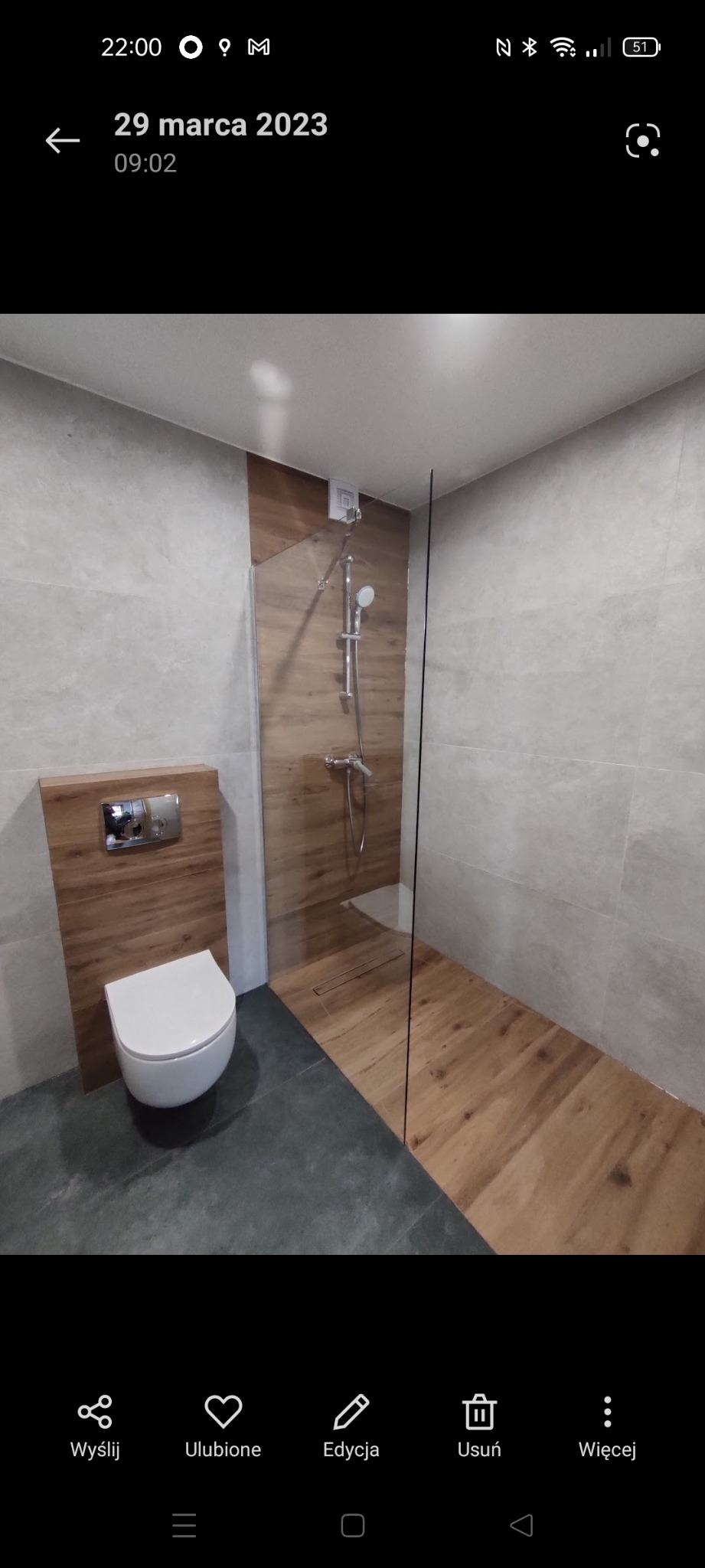 Nowoczesna łazienka z kabiną prysznicową typu walk-in, drewnopodobnymi płytkami i podwieszaną toaletą. Szare płytki na ścianach i podłodze.