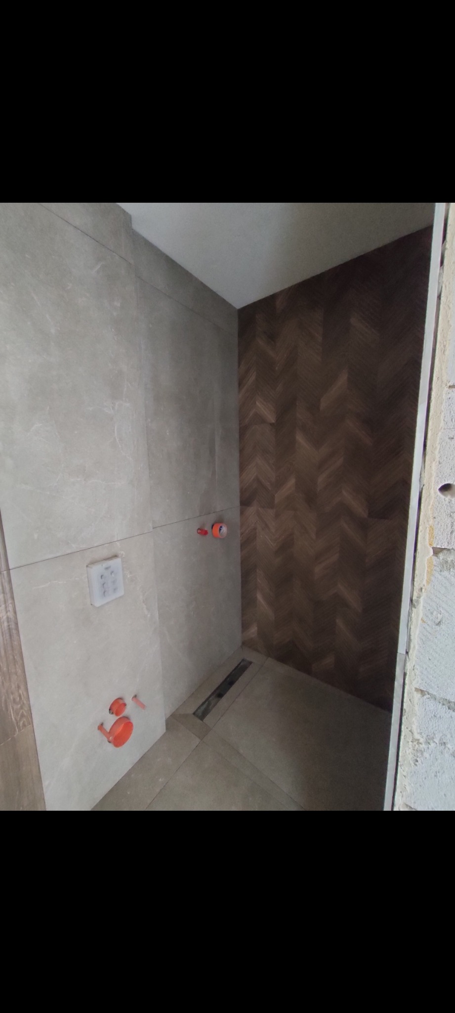 Łazienka w trakcie remontu: szare płytki marmurowe na ścianie z instalacją, brązowe płytki w jodełkę na drugiej ścianie, odpływ liniowy w podłodze.