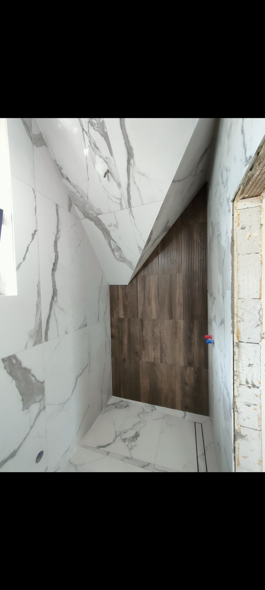 Ukończona łazienka na poddaszu: białe płytki imitujące marmur na ścianach i podłodze, brązowe płytki drewnopodobne pod skosem. Widoczne instalacje wodne.