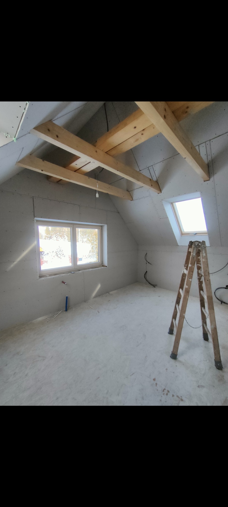 Poddasze w trakcie adaptacji: ściany obłożone płytami gips-karton, widoczne drewniane belki stropowe, okno dachowe i drabina. Prace wykończeniowe.