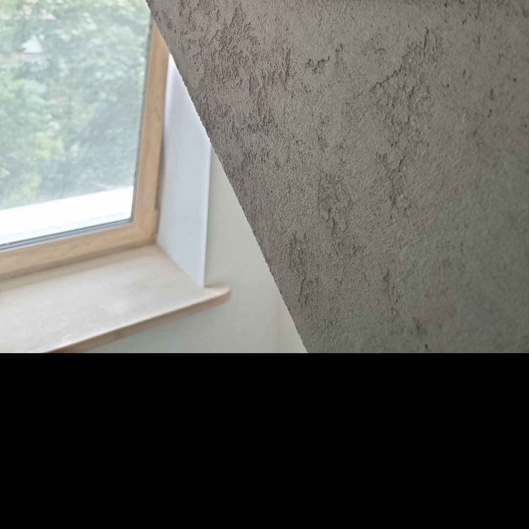 Szara ściana ze strukturą przy oknie dachowym z drewnianą ramą i parapetem, widok z dołu, blisko okna, nierówna faktura.