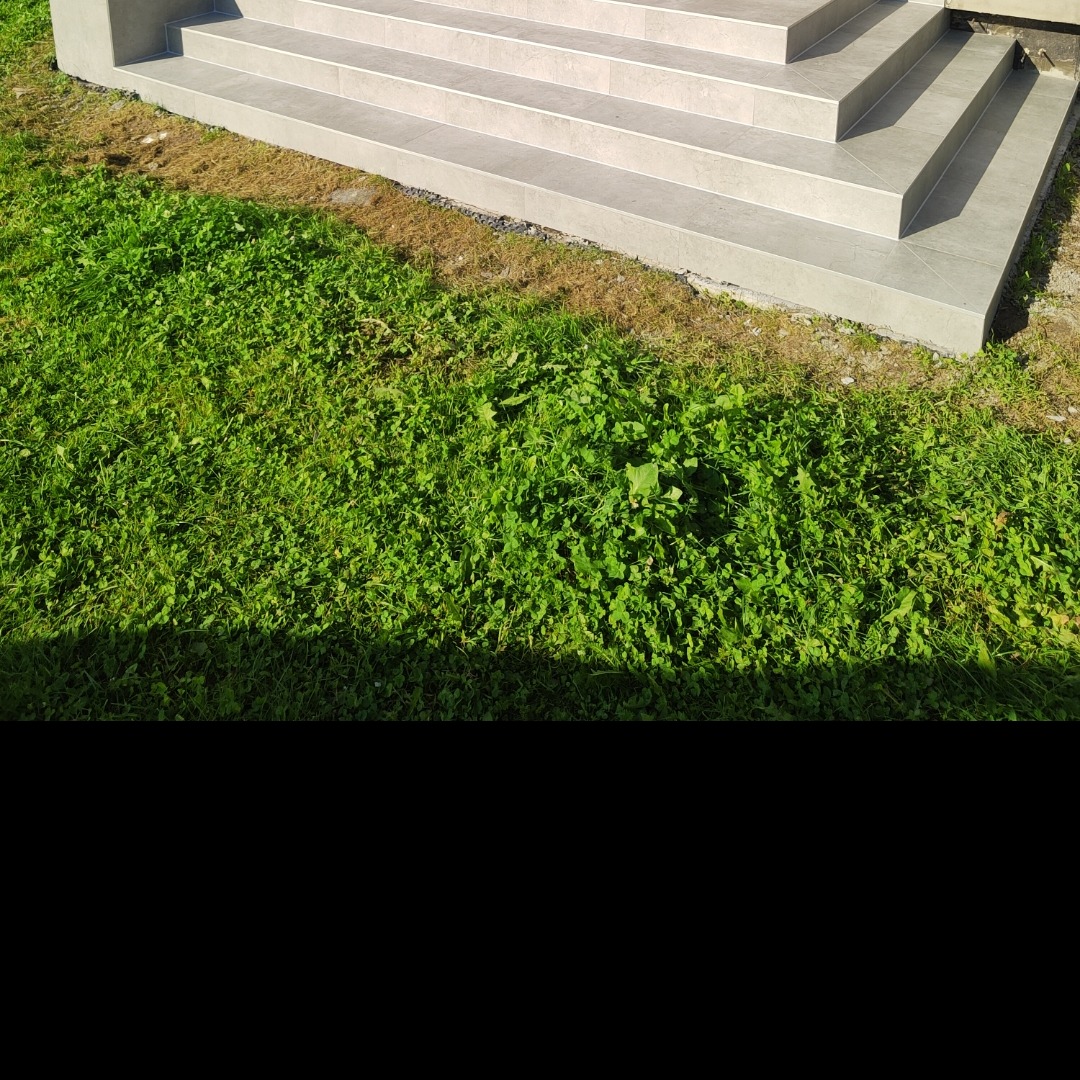 Zewnętrzne schody wyłożone jasnoszarymi płytkami, z widoczną zieloną trawą i fragmentem elewacji budynku. Kąt ujęcia z dołu podkreśla solidność wykonania.