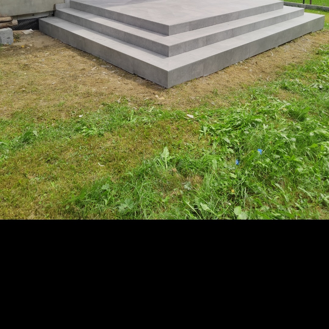 Zewnętrzne schody z szarych płyt, prowadzące do budynku. Widoczne trzy stopnie na tle trawy i fragmentu elewacji. Minimalistyczny design, świeże wykończenie.