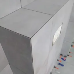 Szczegółowy widok na narożnik obudowy z szarych płytek ceramicznych w trakcie montażu, z użyciem systemu poziomowania płytek.