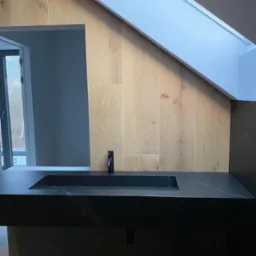 Nowoczesna łazienka na poddaszu: czarna, kamienna umywalka z minimalistyczną baterią, ściana wyłożona płytkami imitującymi drewno, okno dachowe w skosie.