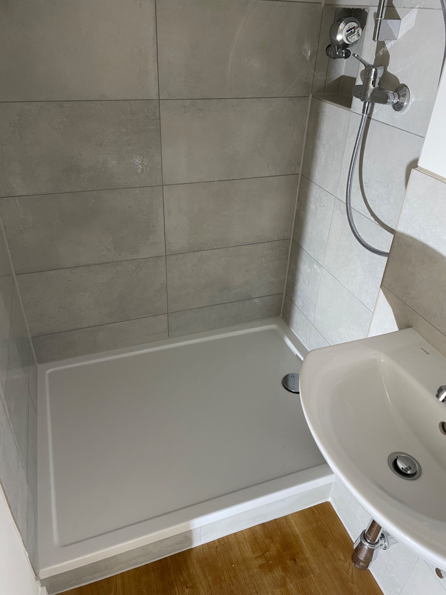 Nowoczesna łazienka z szarymi płytkami, białą kabiną prysznicową i umywalką z chromowaną armaturą, widoczna drewnopodobna podłoga.