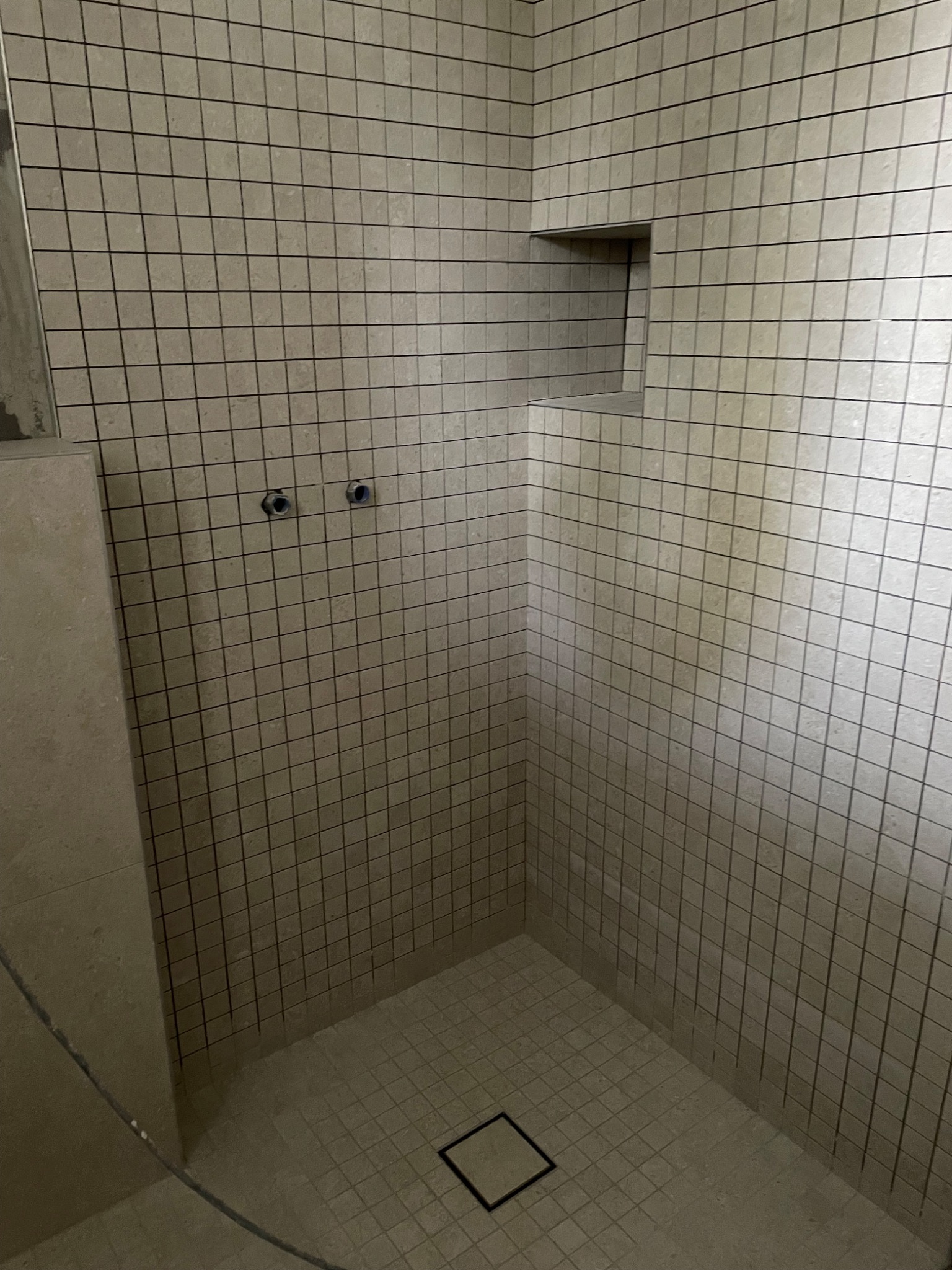 Wnętrze kabiny prysznicowej z beżowymi, kwadratowymi płytkami, widoczne przyłącza wodne i wnęka w ścianie.