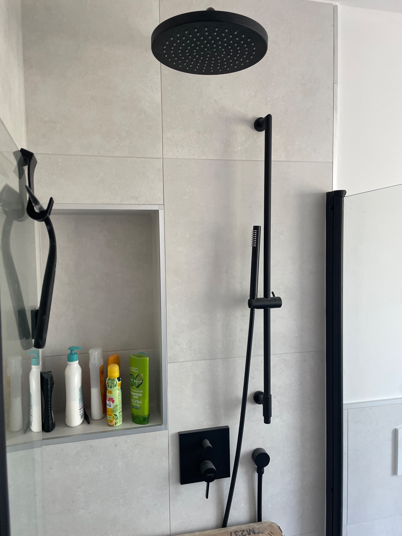 Nowoczesna kabina prysznicowa z czarną armaturą, deszczownicą i wnęką z kosmetykami na ścianie wyłożonej jasnymi płytkami.