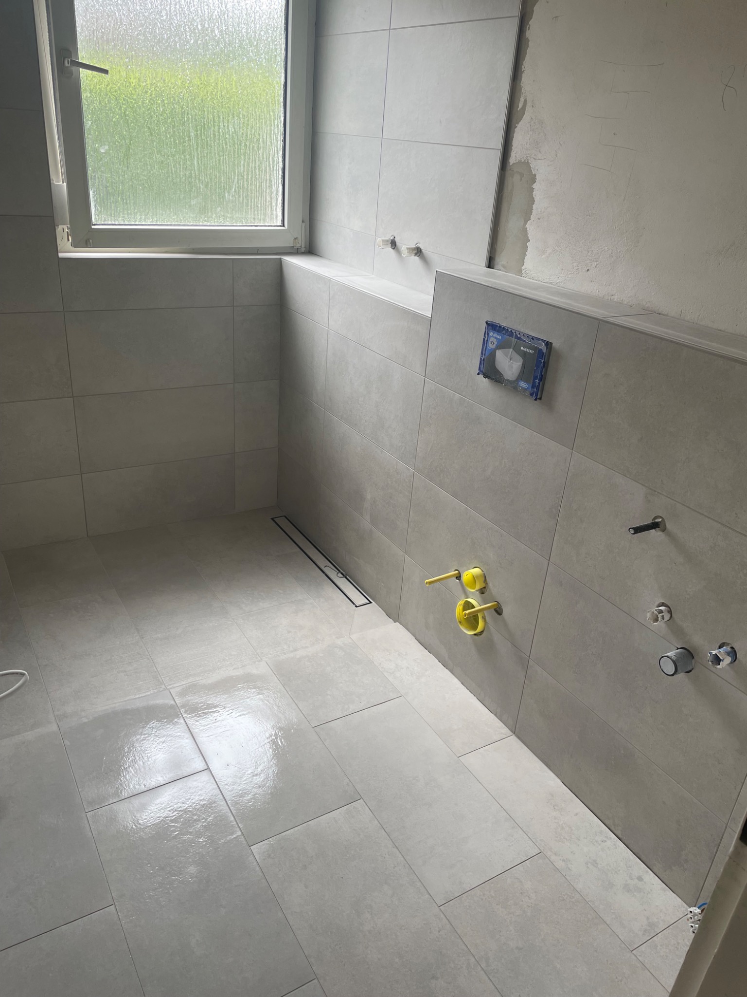 Jasna łazienka w trakcie wykańczania z widocznym odpływem liniowym, częściowo położonymi płytkami i przygotowanymi instalacjami wodno-kanalizacyjnymi, okno z widokiem na zieleń.