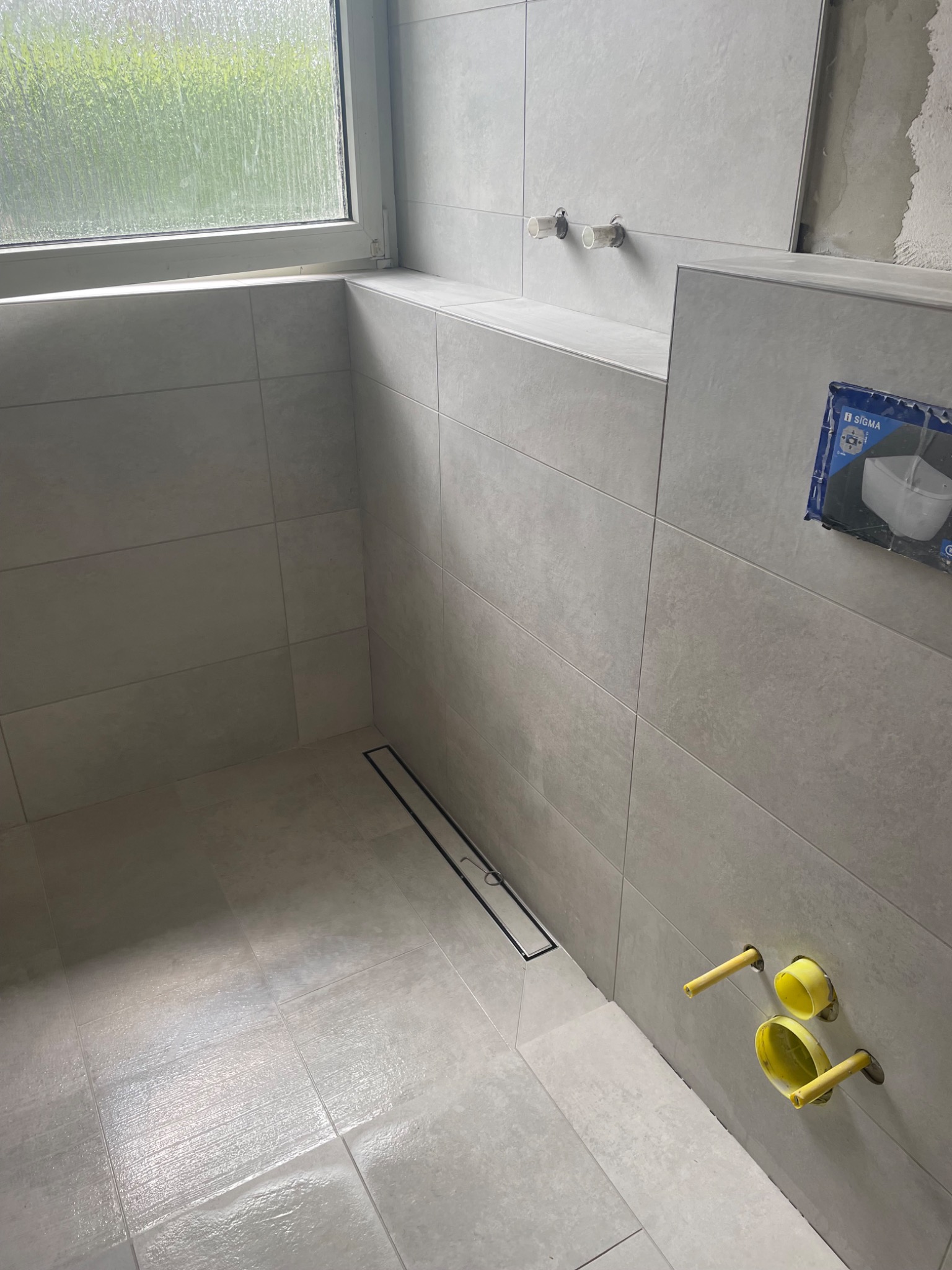 Łazienka w trakcie remontu: szare płytki na ścianach i podłodze, zamontowany odpływ liniowy, wystające rury z żółtymi zaślepkami, widok na zewnątrz przez okno.