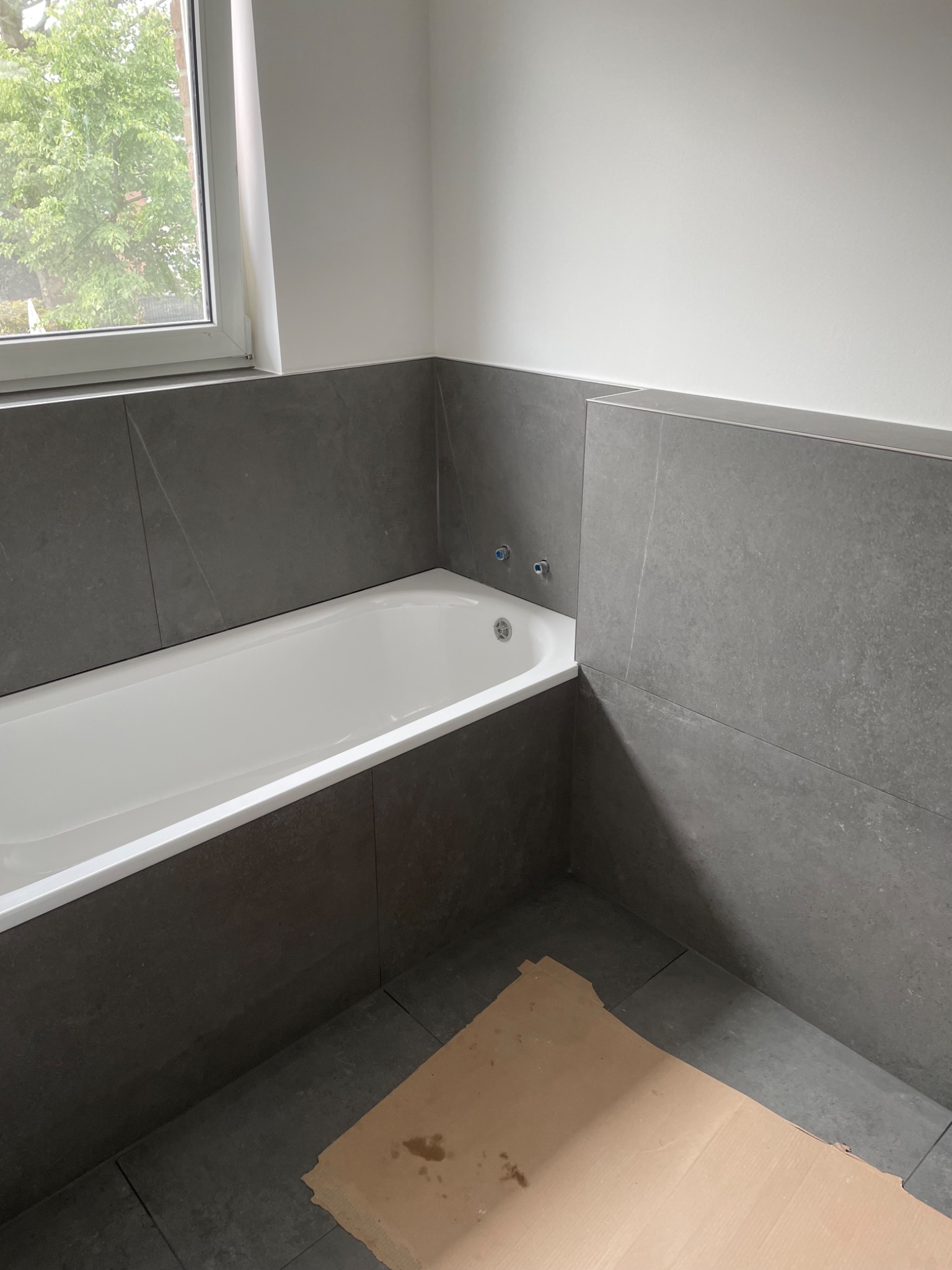 Wanna obudowana szarymi płytkami, widoczne dwa zawory na ścianie, podłoga wyłożona płytkami w odcieniach szarości, zabezpieczona kartonem.