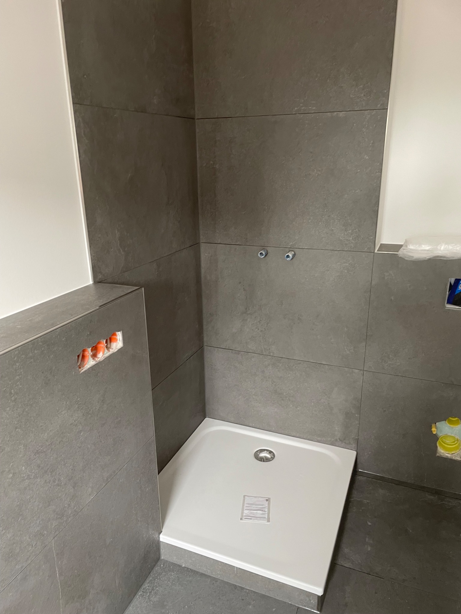 Biały brodzik w rogu łazienki wyłożonej szarymi, matowymi płytkami; widoczne otwory na baterię i instalację wodną.