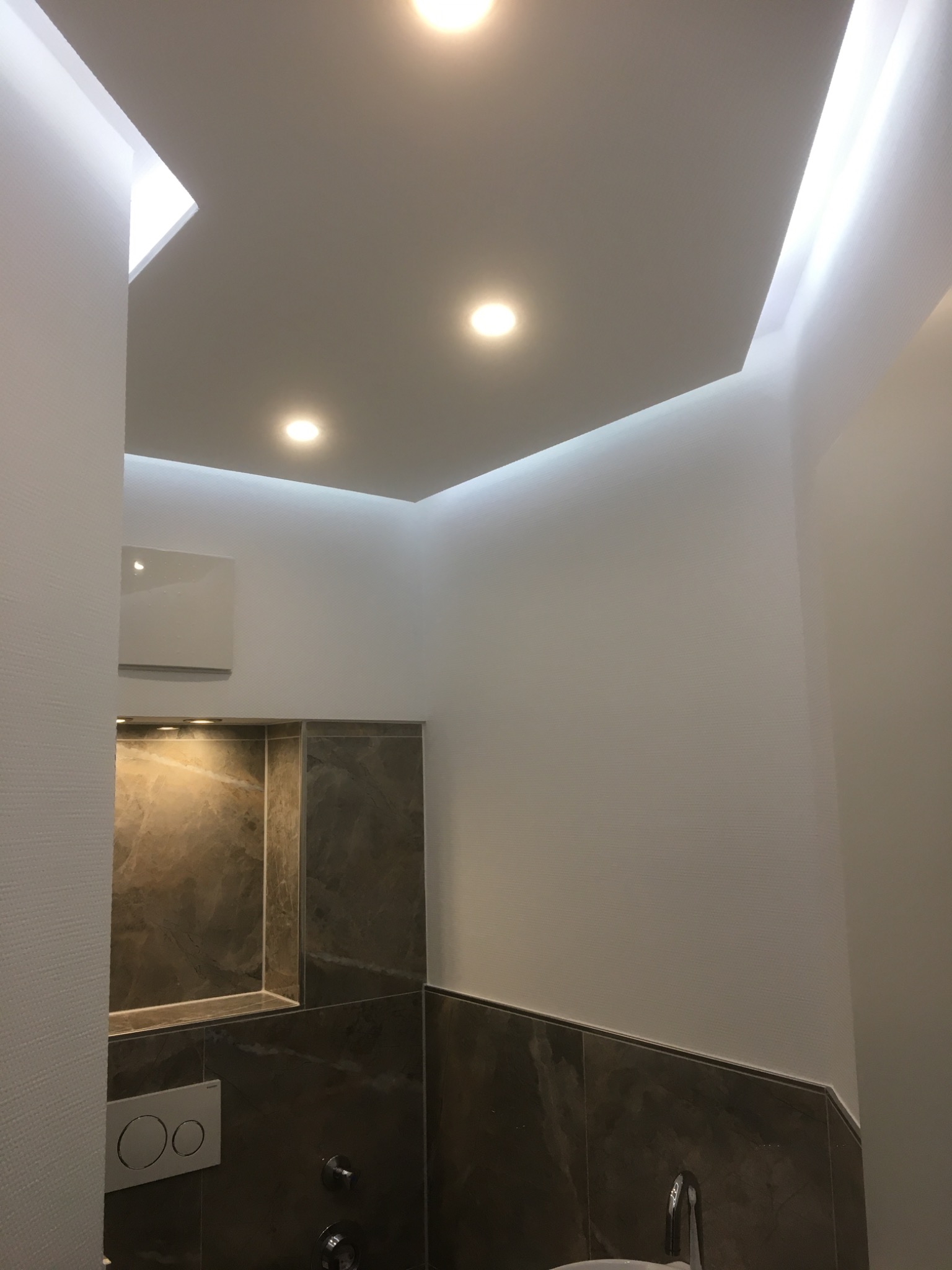 Nowoczesna łazienka z podświetlanym sufitem, ciemnymi płytkami imitującymi marmur, wiszącą miską WC i wnęką z oświetleniem.