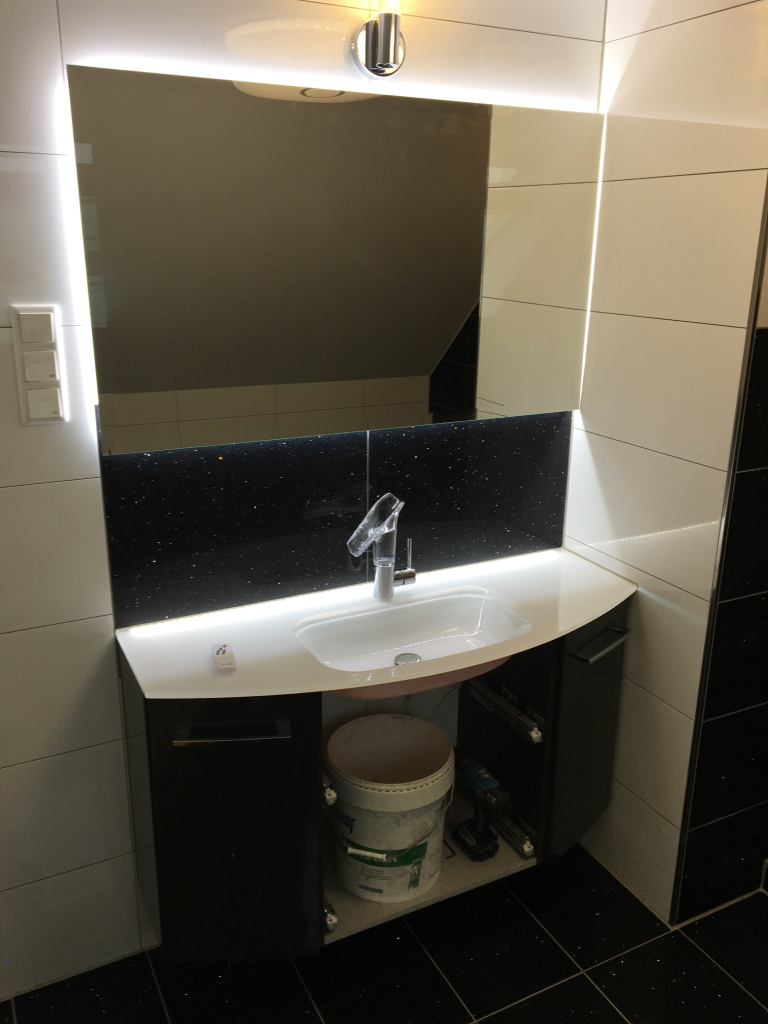 Nowoczesna łazienka z podświetlanym lustrem i czarnym brokatowym panelem nad umywalką, widoczny biały blat, bateria umywalkowa, biała umywalka, biało-czarne płytki na ścianach, wiadro i wiertarka...