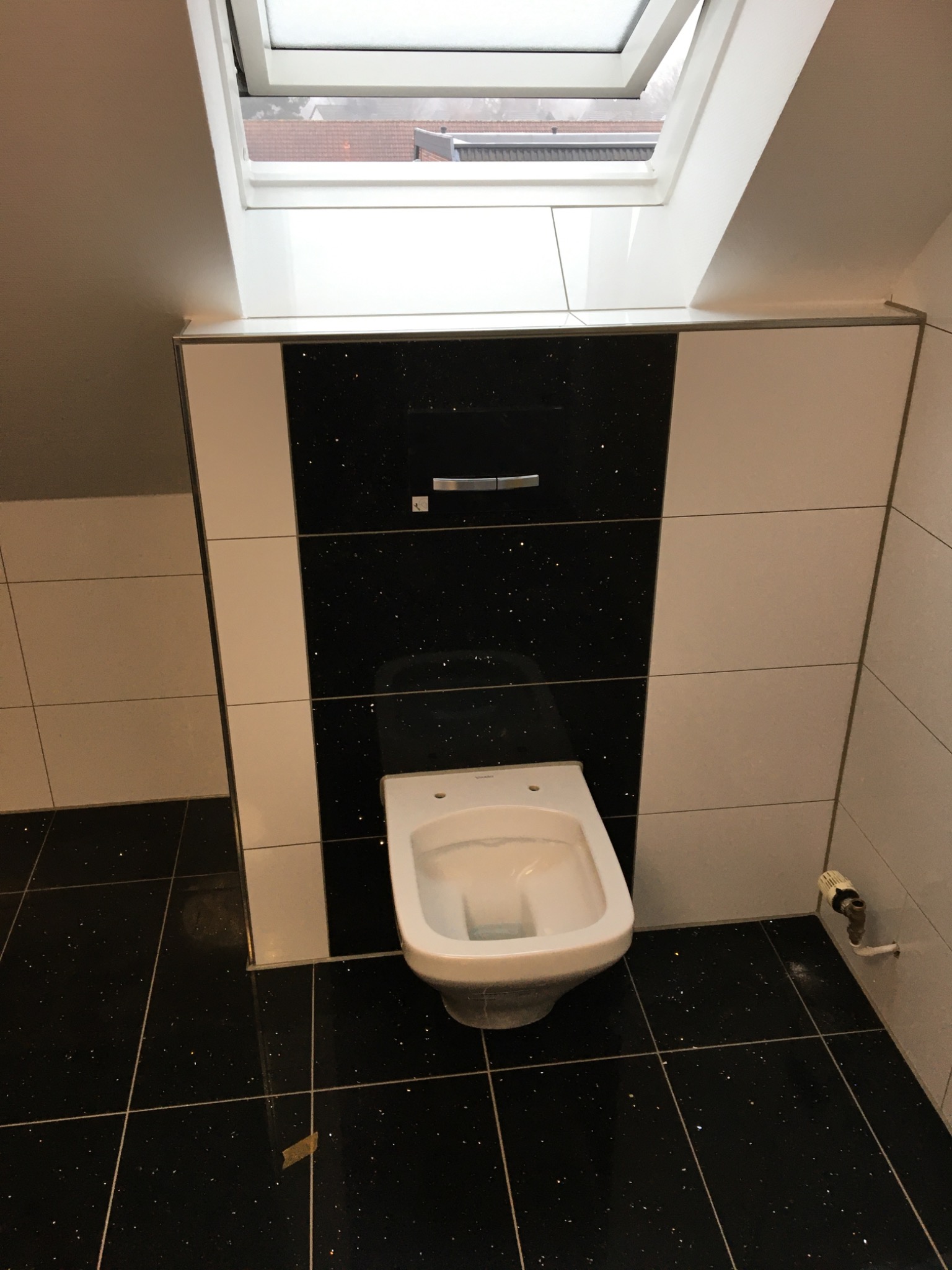 Nowoczesna toaleta podwieszana w łazience na poddaszu, z czarno-białymi płytkami z brokatem, widok na okno dachowe.