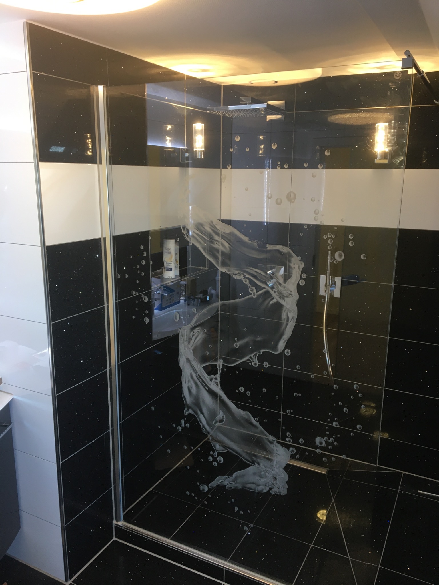 Szklana kabina prysznicowa z czarnymi, błyszczącymi płytkami i białymi akcentami, pokryta kroplami wody po prysznicu, widoczne oświetlenie sufitowe i dozownik mydła.