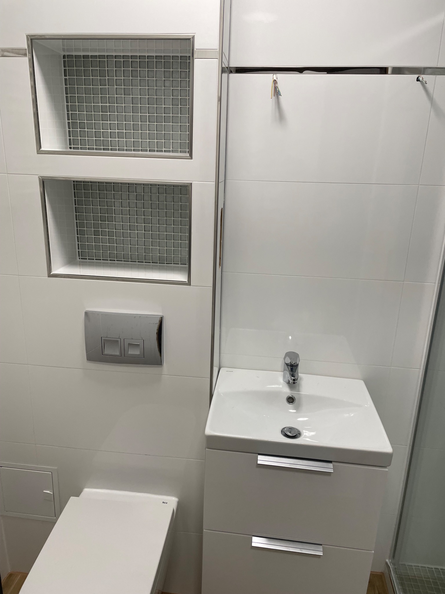 Nowoczesna łazienka wykończona białymi płytkami z dwoma wnękami z mozaiką, wisząca szafka z umywalką i toaleta.