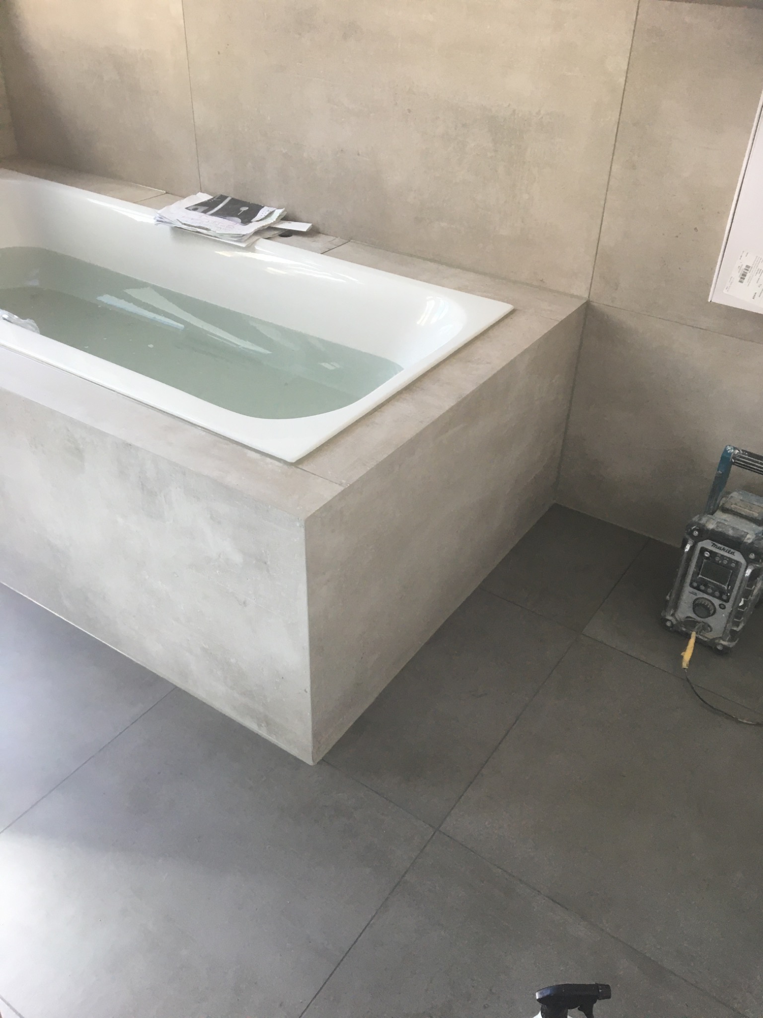 Wanna wypełniona wodą, obudowana płytkami imitującymi beton, na wierzchu leżą projekty, na podłodze płytki w odcieniach szarości, widoczny sprzęt budowlany.