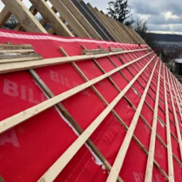 Konstrukcja drewniana dachu pokryta czerwoną membraną, widoczne elementy więźby dachowej i fragmenty łat, w tle zabudowa jednorodzinna i pochmurne niebo.