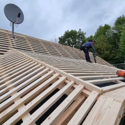 Ryszard Dekarz Robo Dach - Montaż drewnianej konstrukcji dachu z użyciem narzędzi pneumatycznych, widoczny pracownik w trakcie mocowania łat na folii dachowej, w tle drzewa i antena satelitarna.