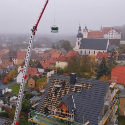 Ryszard Dekarz Robo Dach - Montaż nowego dachu z wykorzystaniem dźwigu na tle zabudowy miejskiej z kościołem w tle, widoczne rusztowania i częściowo ułożona dachówka. Dach pokryty szarą dachówką.