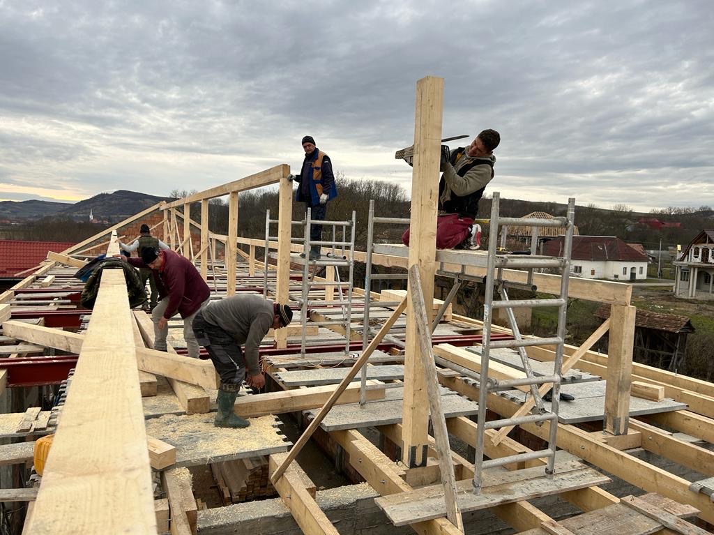 Ekipa budowlana montuje drewnianą konstrukcję dachu na tle pochmurnego nieba i zabudowań wiejskich. Jeden z pracowników, siedząc na rusztowaniu, przycina drewniany słup piłą elektryczną.