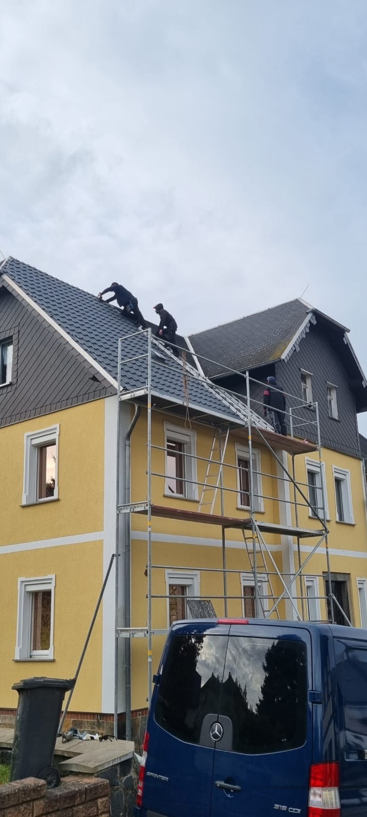 Ekipa dekarzy na rusztowaniu podczas wymiany pokrycia dachu z łupka na żółtym budynku mieszkalnym, widoczny niebieski samochód dostawczy Mercedes Sprinter.