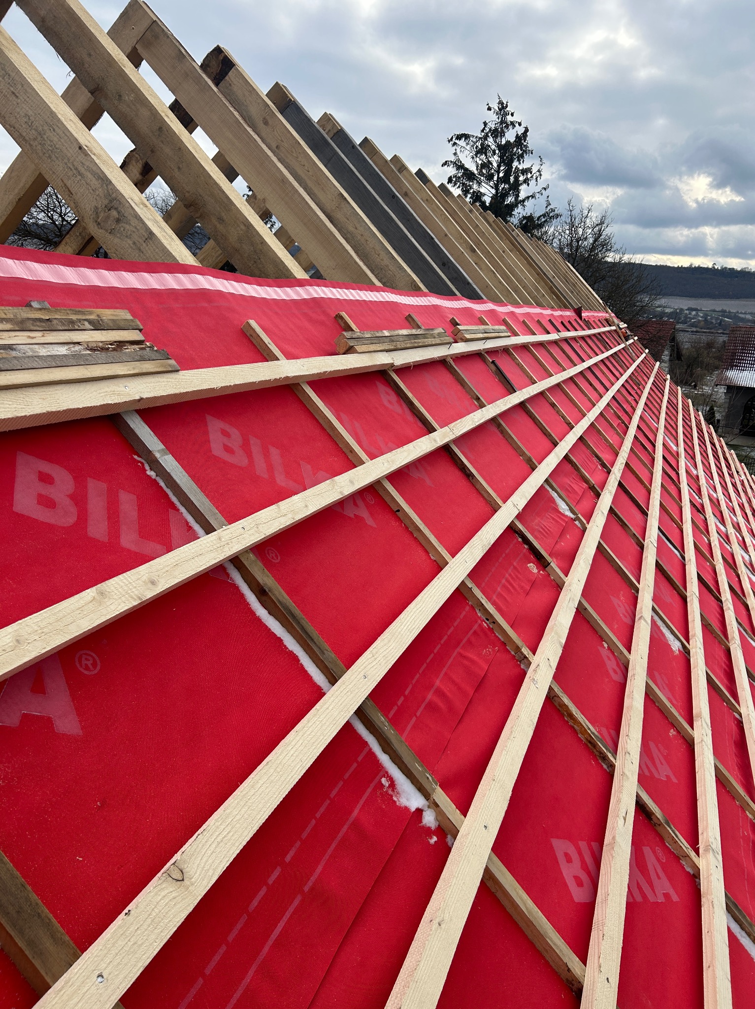Konstrukcja drewniana dachu pokryta czerwoną membraną, widoczne elementy więźby dachowej i fragmenty łat, w tle zabudowa jednorodzinna i pochmurne niebo.