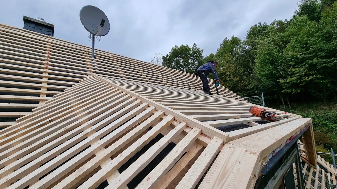 Montaż drewnianej konstrukcji dachu z użyciem narzędzi pneumatycznych, widoczny pracownik w trakcie mocowania łat na folii dachowej, w tle drzewa i antena satelitarna.