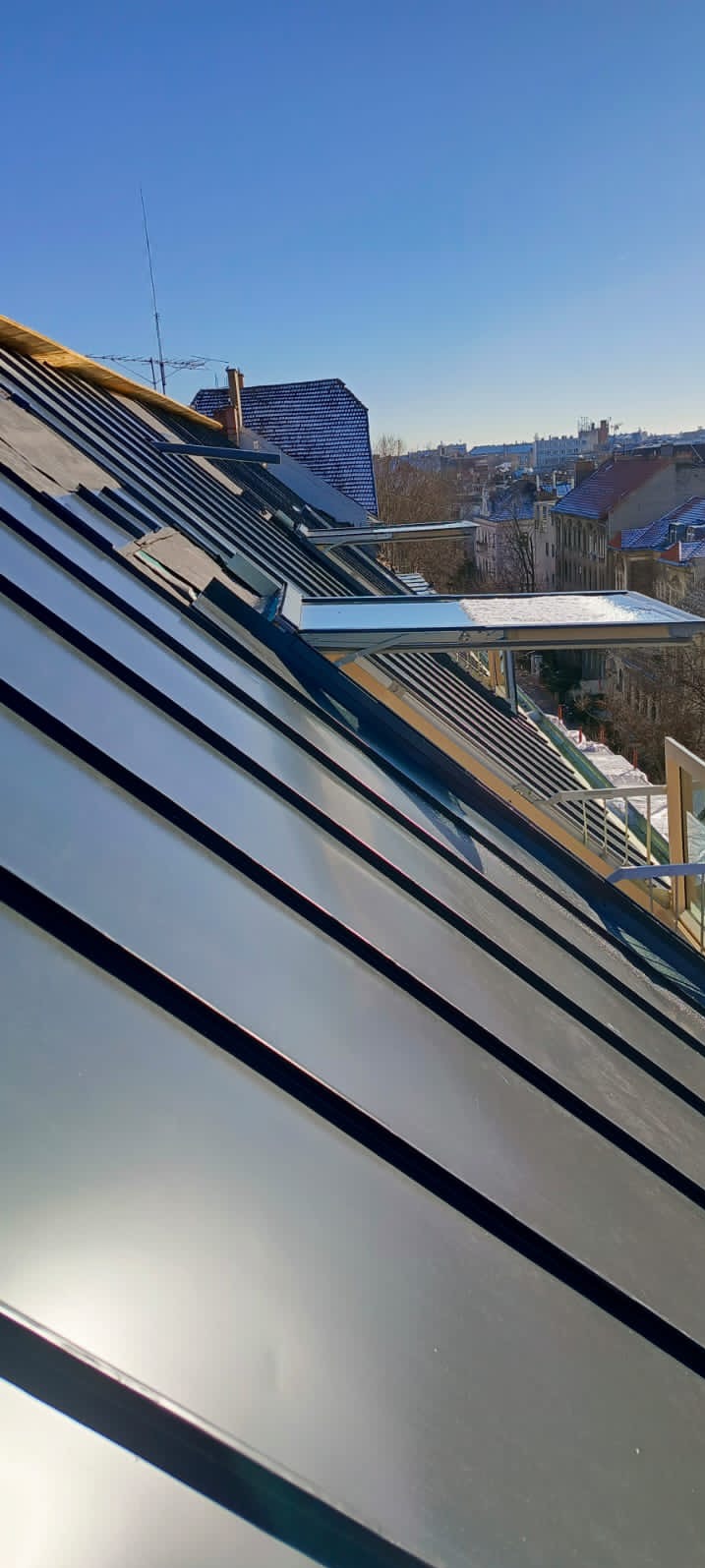 Nowy, błyszczący, grafitowy dach z blachy na tle błękitnego nieba i miejskiej zabudowy, widoczne okna dachowe.