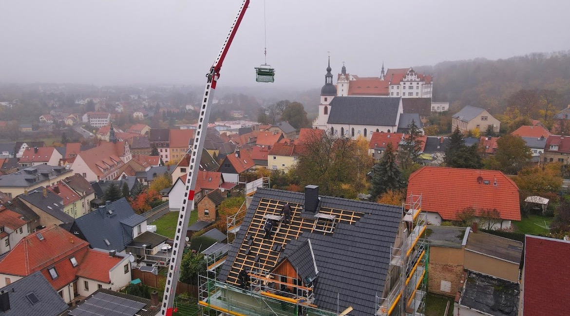 Montaż nowego dachu z wykorzystaniem dźwigu na tle zabudowy miejskiej z kościołem w tle, widoczne rusztowania i częściowo ułożona dachówka. Dach pokryty szarą dachówką.