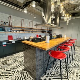 PRIMATER SPÓŁKA Z OGRANICZONĄ ODPOWIEDZIALNOŚCIĄ - Nowoczesna kuchnia biurowa z wyspą, drewnianym blatem, czerwonymi hokerami i wzorzystą podłogą w czarno-białe geometryczne wzory. Widoczne półki z naczyniami i ekspres do kawy.